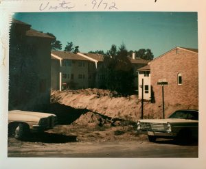 Photo of deep holes in ground with mounded dirt, surrounded by some other apartment buildings and the front and back of two old cars