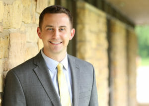 Head shot of Derek Madsen. He's wearing a gray suit coat, light blue shirt, and yellow tie, and smiling at the camera.