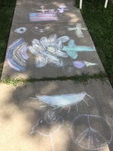 This image shows sidewalk chalk art doodles. In the top of the frame is an American flag; just below is an outline of a cat's face. Below that is a rainbow and a large flower. At the bottom of the frame is an upside down sunset or sunrise, an outline of a bird, and a peace sign.